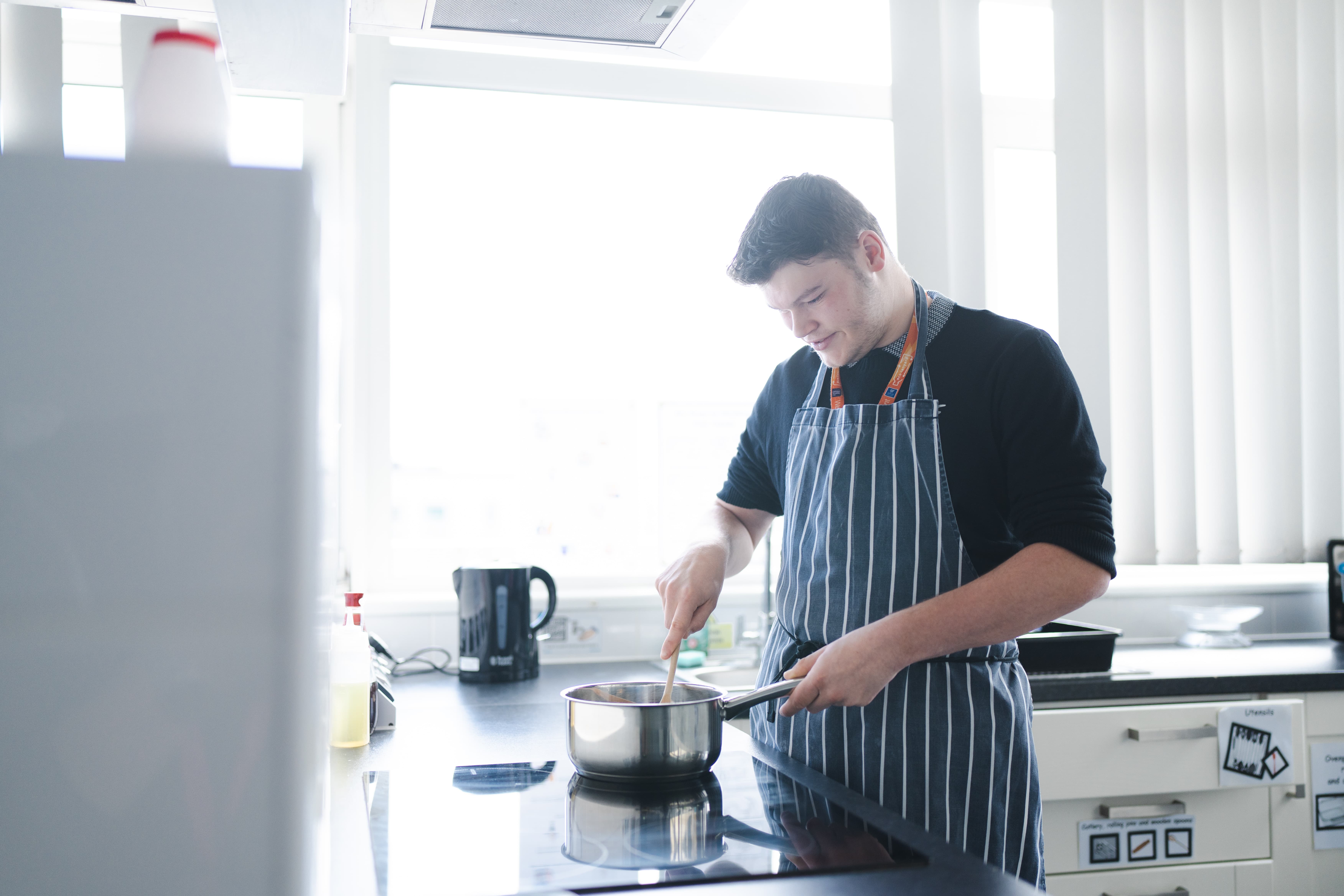 boy standing in a teaching kitchen standing over a stove stirring something in a saucepan