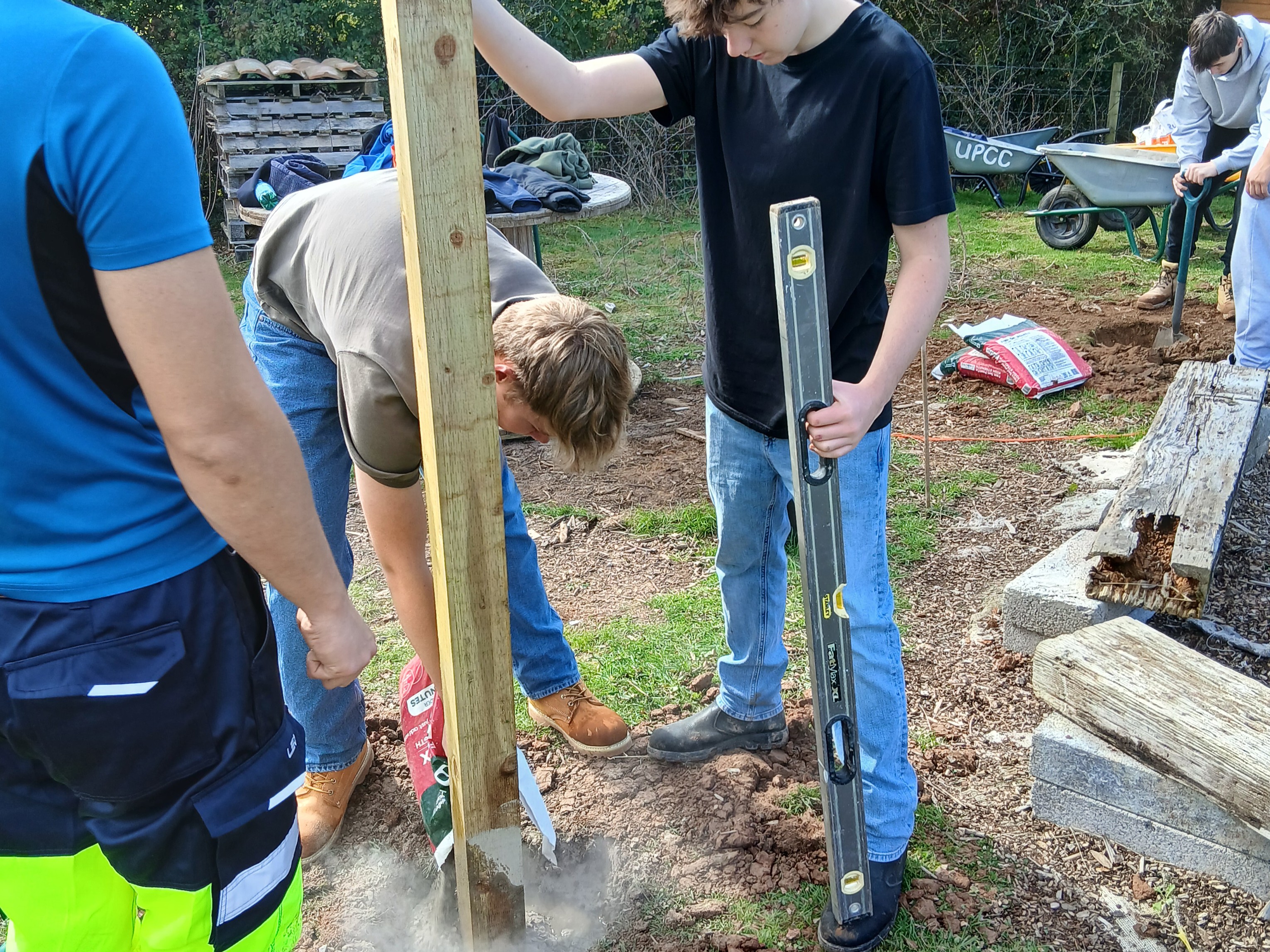 Student holding the wooden post upright in the ground and putting cement into the hole