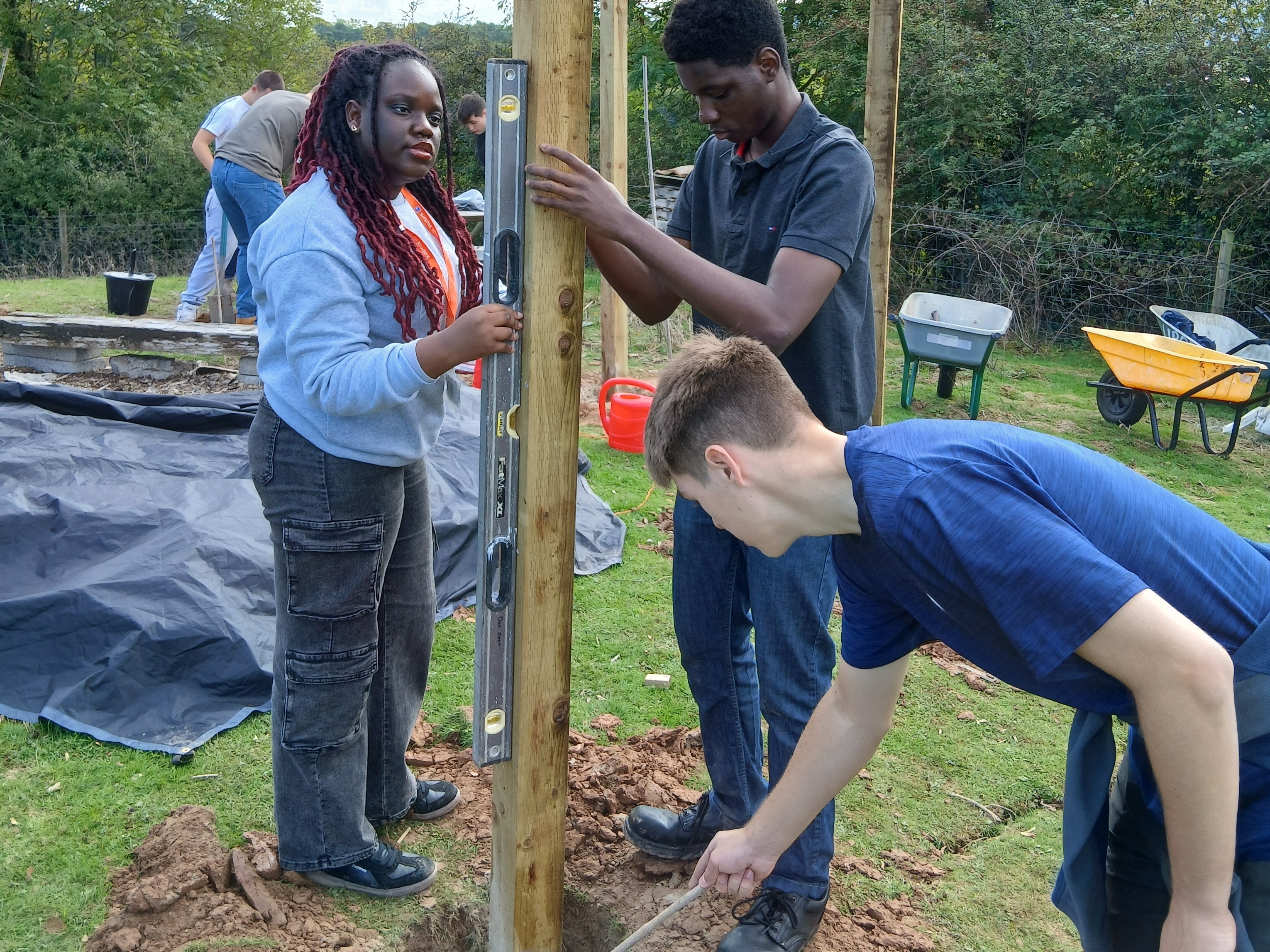 Student holding the wooden post upright in the ground and putting cement into the hole