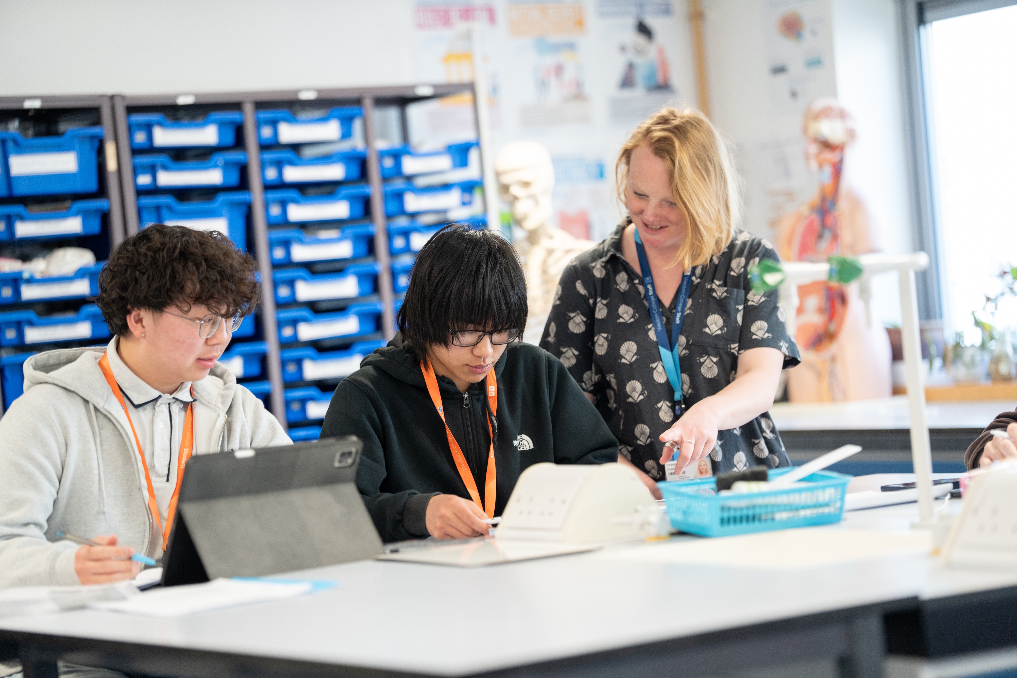 two boys sat at a desk on computers, with a teacher smiling over them