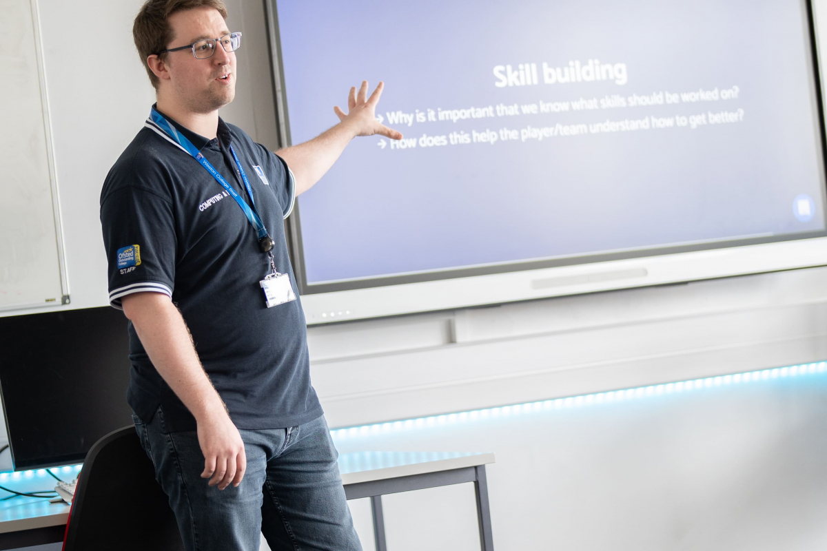 lecturer at the front of a classroom pointing to an interactive whiteboard 