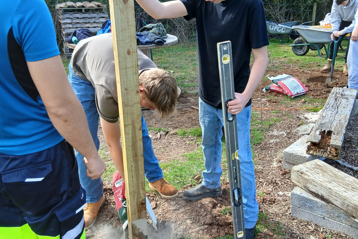 Student holding the wooden post upright in the ground and putting cement into the hole 
