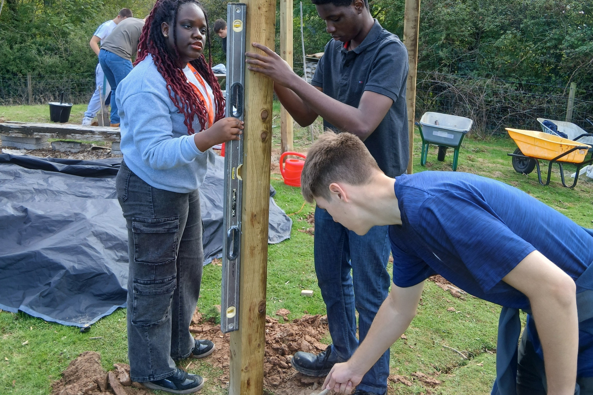 Student holding the wooden post upright in the ground and putting cement into the hole 