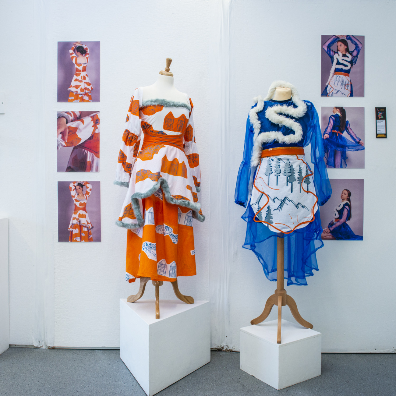 Students work on show in art exhibition at the summer show. One orange dress and one blue dress with fur trim on mannequins with photographs of a model wearing the garments on the wall behind