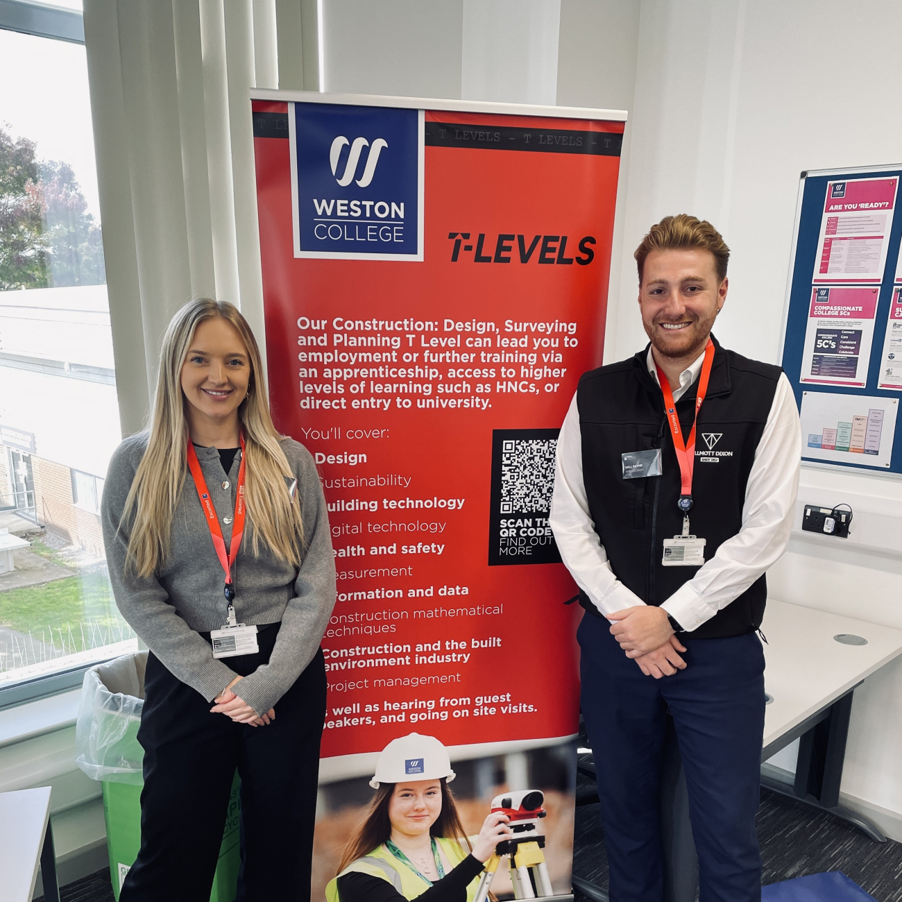 Will and Chloe from Willmott Dixon stood in front of a T Level banner smiling